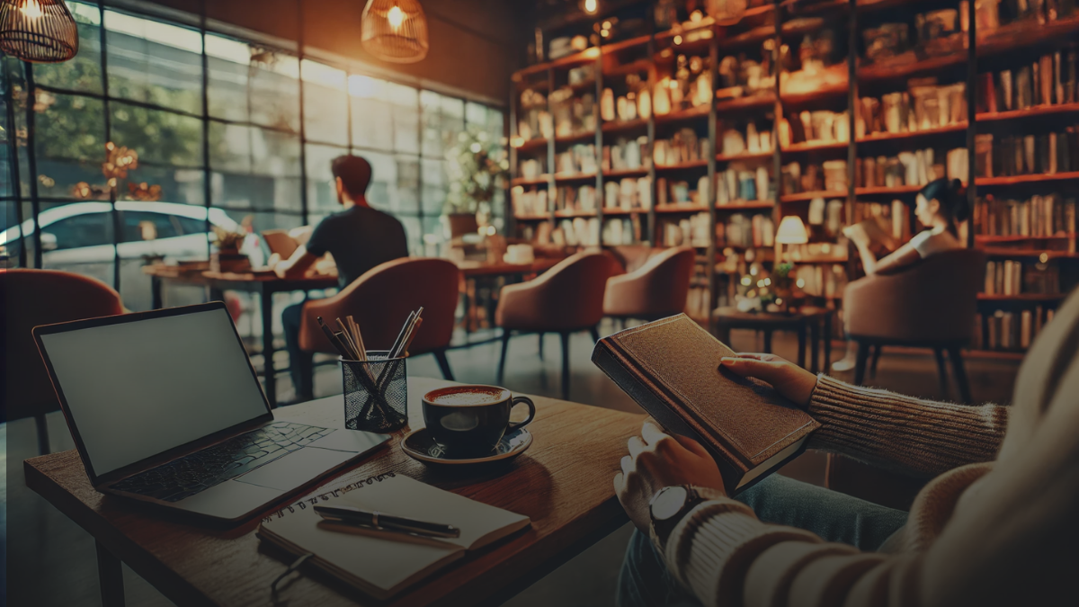 Person reading a book in a cozy coffee shop with a notebook and laptop nearby.